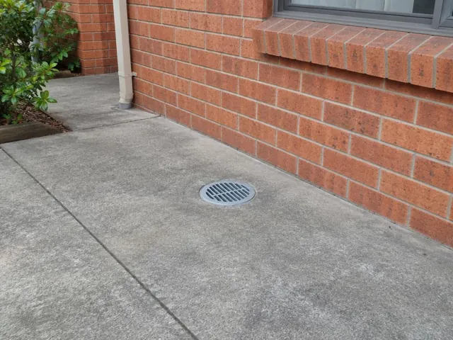 Dry concrete path with a round drain grate beside a brick house wall in Kirrawee